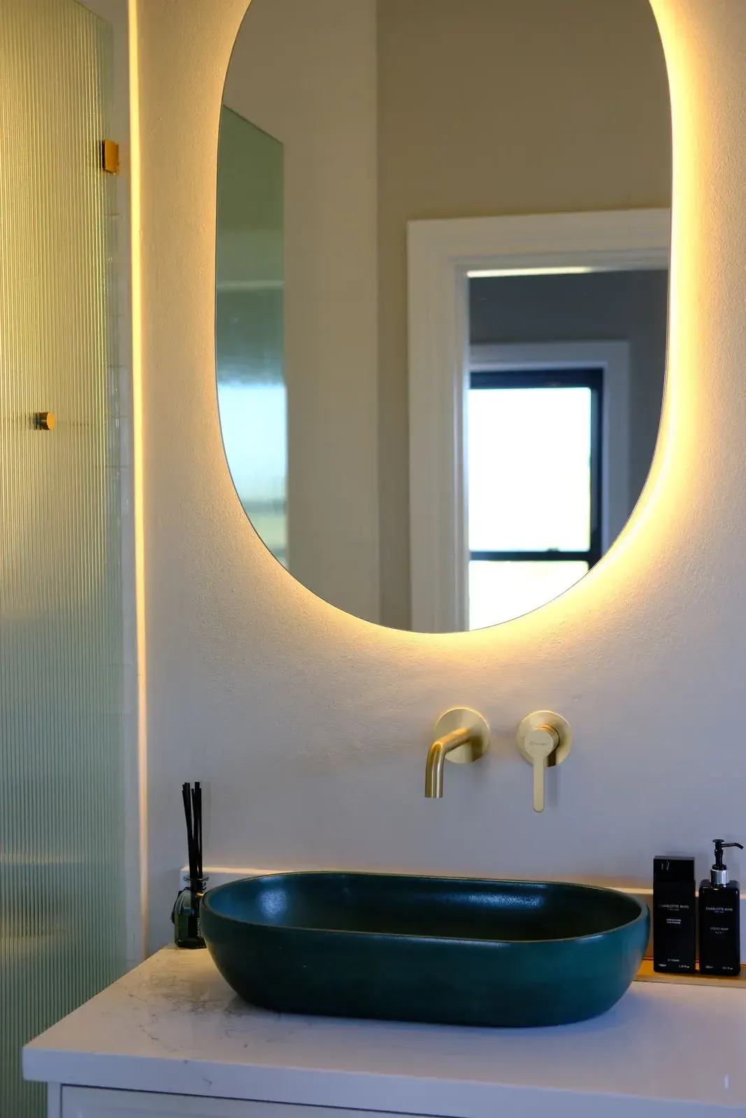 Brushed brass wall-mounted basin mixer with a backlit mirror and teal basin