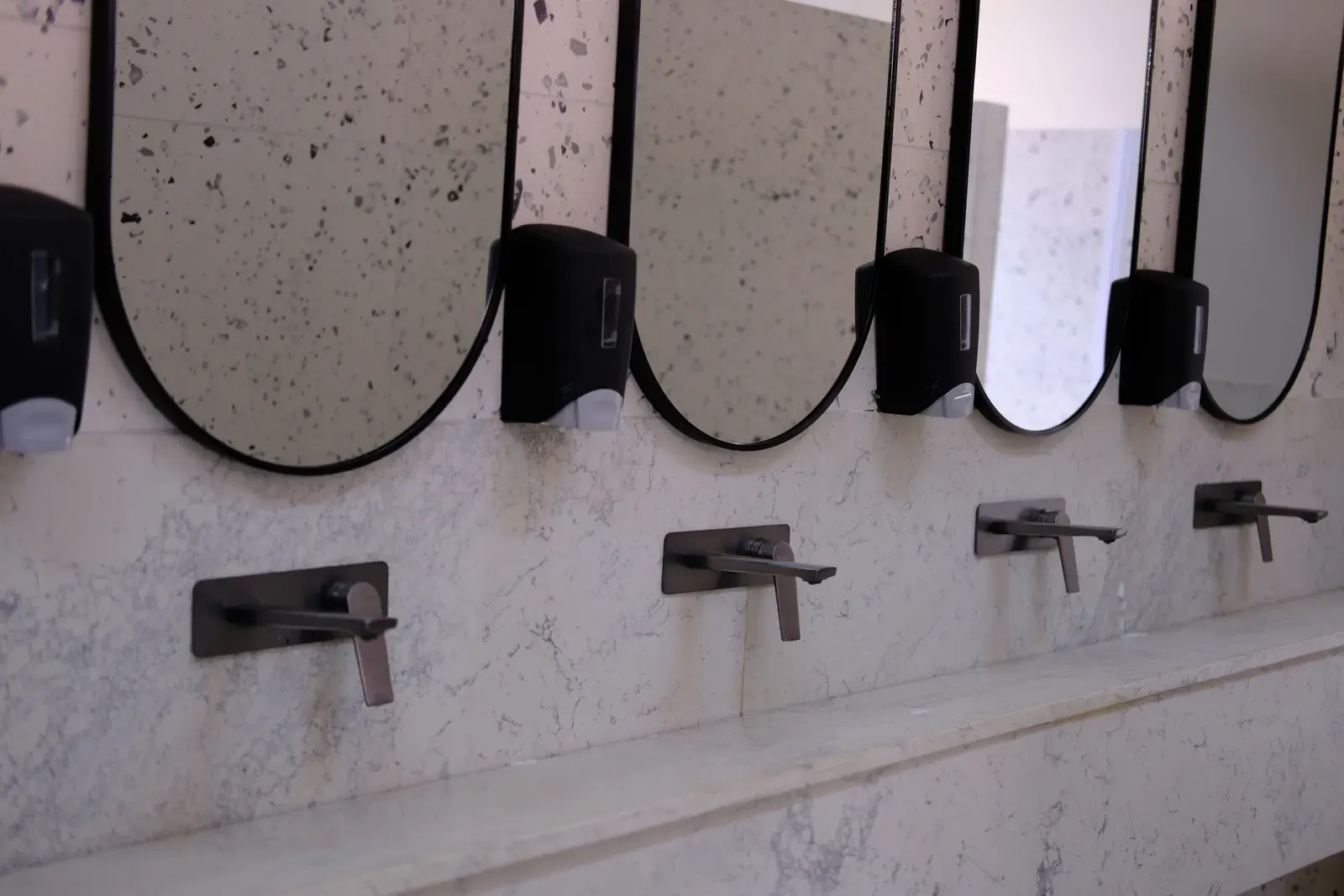 Row of matte black wall-mounted basin mixers under arched mirrors