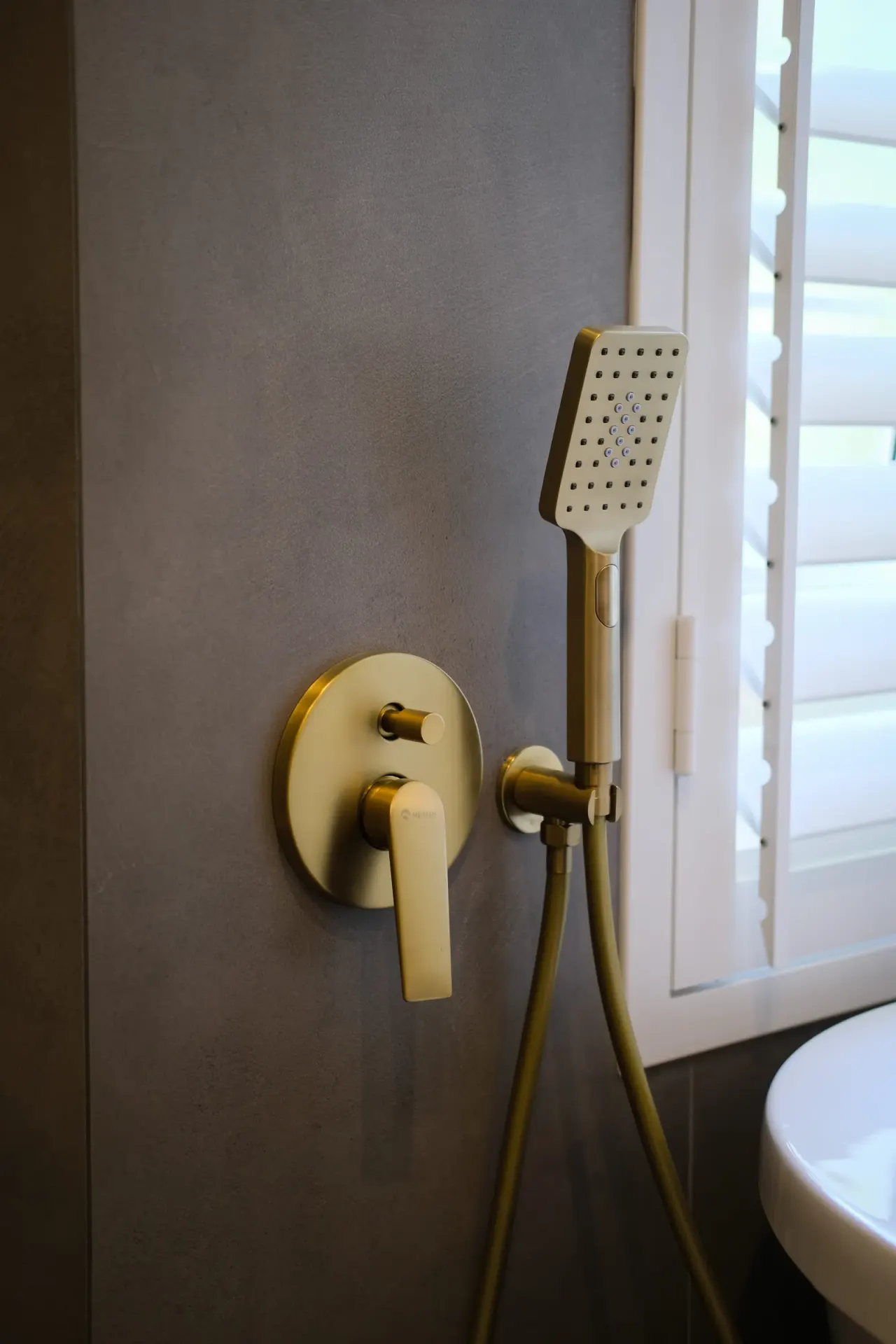 Brushed brass wall-mounted mixer and square handheld shower by a shuttered window