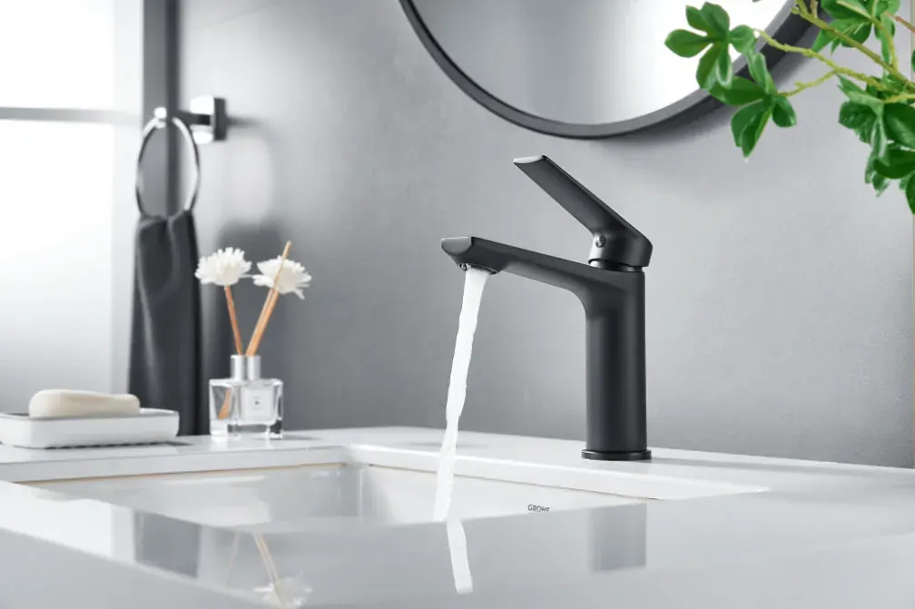 Matte black single-lever basin mixer running water into an undermount basin, with a black towel ring and round mirror