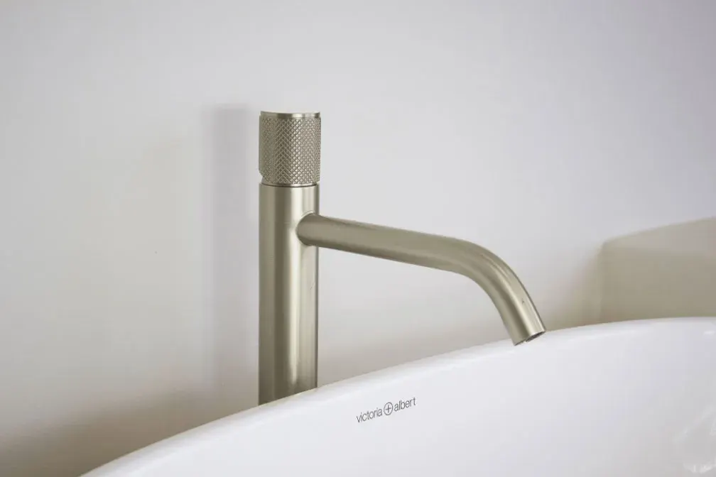 Brushed-finish tall basin mixer with knurled detailing beside a white freestanding bath